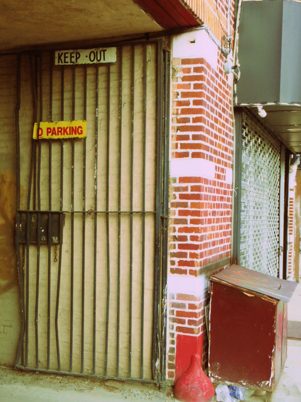 Photo of an open, black barred gate next to a brick wall. Attached signs read "No Parking" and "Keep Out"